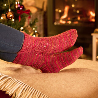 Feet in red knitted socks rest on a blanket by a cosy fireplace, with a decorated Christmas tree in the background, creating a warm, festive atmosphere.