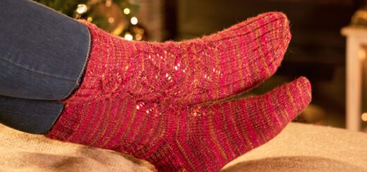 Feet in red knitted socks rest on a blanket by a cosy fireplace, with a decorated Christmas tree in the background, creating a warm, festive atmosphere.