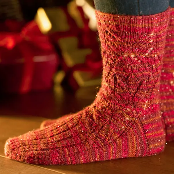 A close-up of vibrant red knitted socks with intricate patterns, worn by a person standing on a wooden floor. Cosy atmosphere with wrapped gifts blurred in the background.