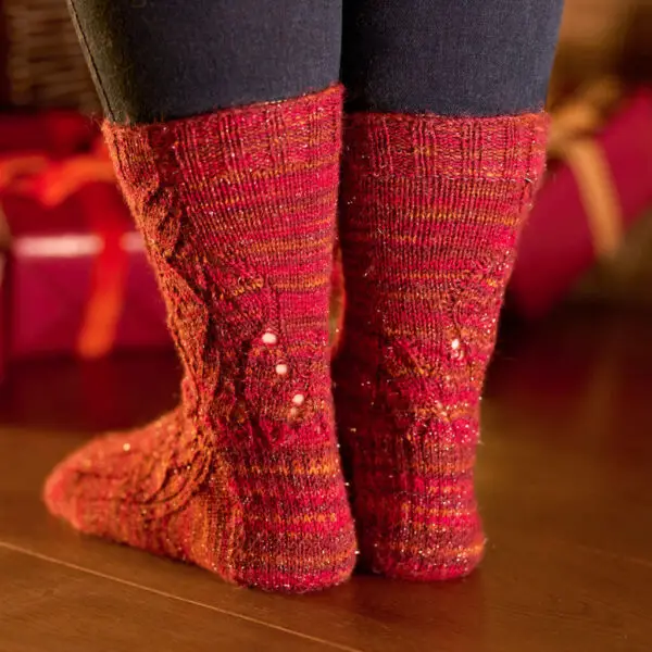 Cozy, festive red knit socks with intricate patterns adorn feet on a wooden floor. A warm, holiday ambiance is enhanced by blurred gift boxes in the background.