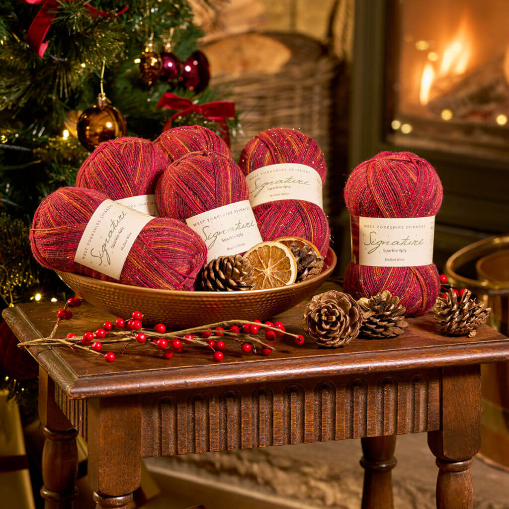 Cosy Christmas scene with red yarn balls in a basket on a wooden table, surrounded by pinecones, dried orange slices, and red berries. A fireplace and tree are in the background, creating a warm and festive atmosphere.