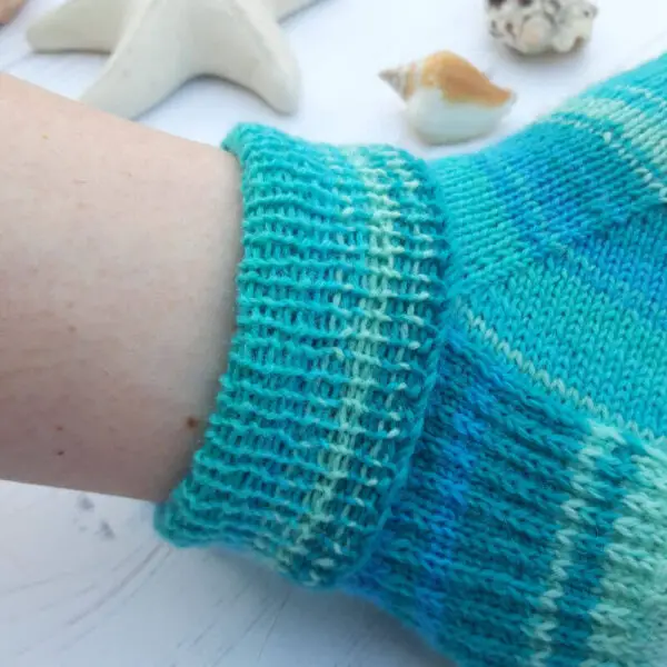 Close-up of a foot wearing a turquoise-striped knitted sock with a ribbed cuff. Background features seashells and a star-shaped ornament on a light surface. Winwick Mum Basic 4ply Shortie Socks