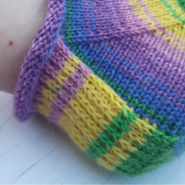 Close-up of a knitted sock heel in vibrant stripes of purple, yellow, green, and blue. Winwick Mum Basic 4ply Shortie Socks