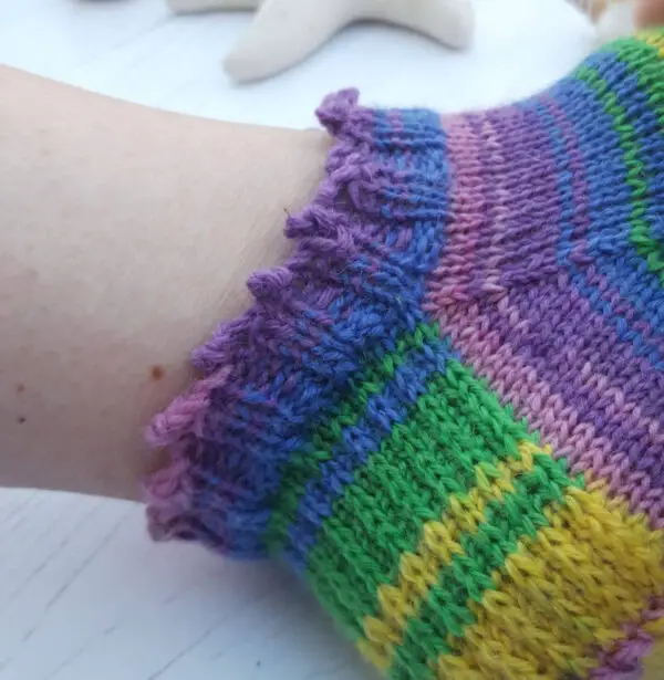 Close-up of a multicoloured knitted sock on an ankle, featuring stripes of purple, pink, green, and yellow with a ruffled picot cuff. Soft, cozy vibe. Winwick Mum Basic 4ply Shortie Socks
