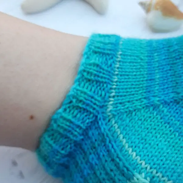 Close-up of a foot wearing a blue-green knitted sock, evoking a cozy feel. Background includes blurred seashells on a white surface. The sock cuff goes straight into the body of the sock. Winwick Mum Basic 4ply Socks