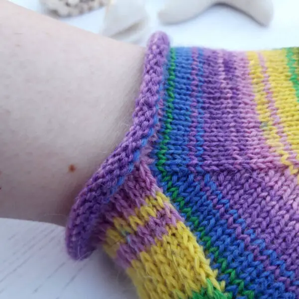 Close-up of a multi-coloured knitted sock on a foot. The sock features vibrant stripes of purple, yellow, blue, and green. The texture appears soft and cozy. The cuff is rolled before joining with the foot. Winwick Mum Basic 4ply Shortie Socks