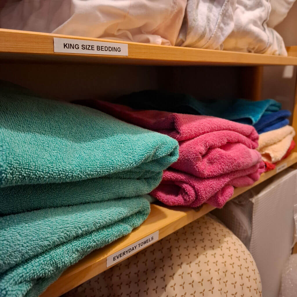 Shelves with neatly folded towels and bedding. Top shelf labeled "King Size Bedding" with white linens; bottom shelf "Everyday Towels" with colourful towels.