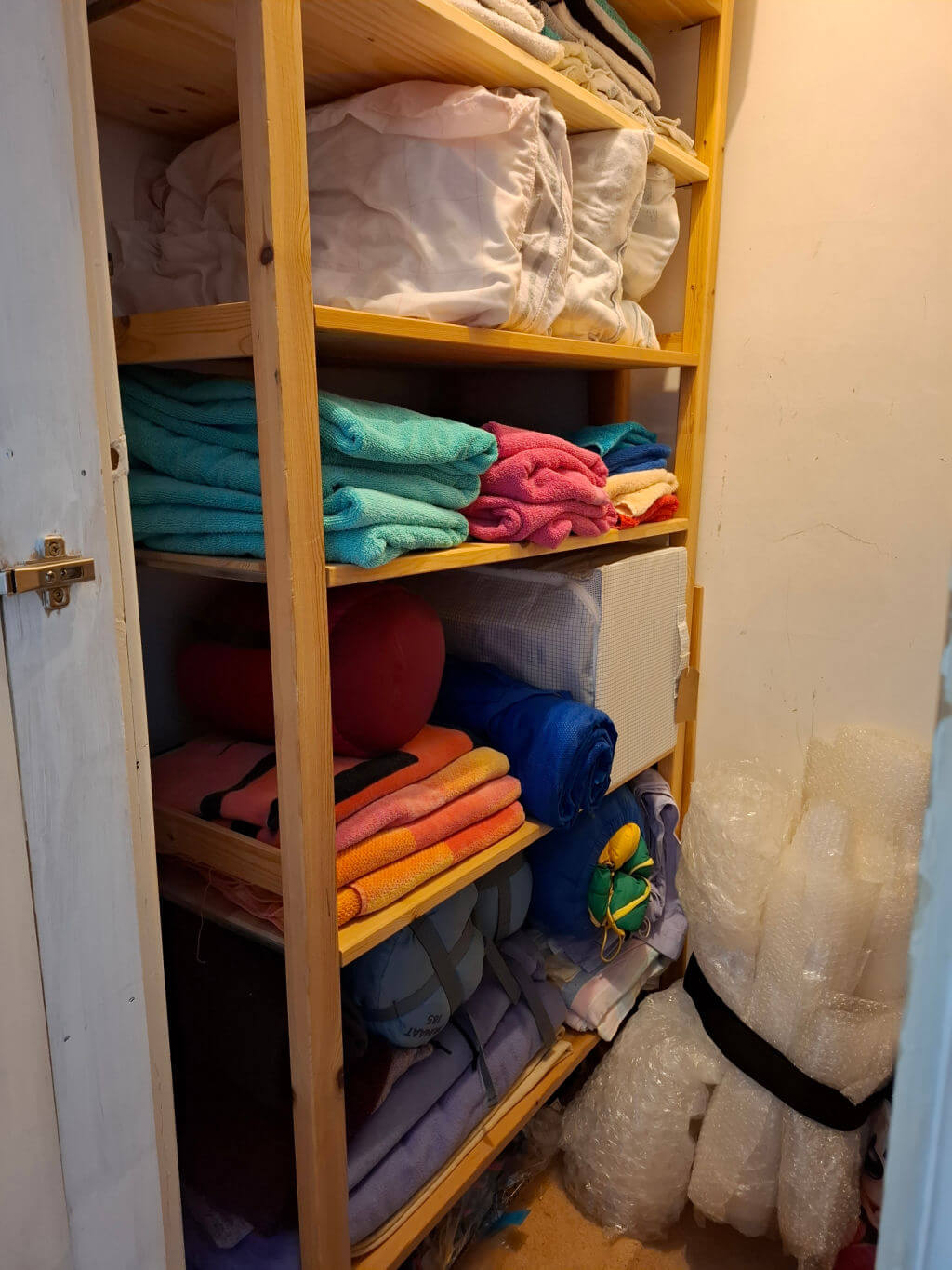 Wooden shelving unit in a closet, neatly stacked with colourful towels, folded sheets, storage boxes, and bubble wrap, creating an organized feel.