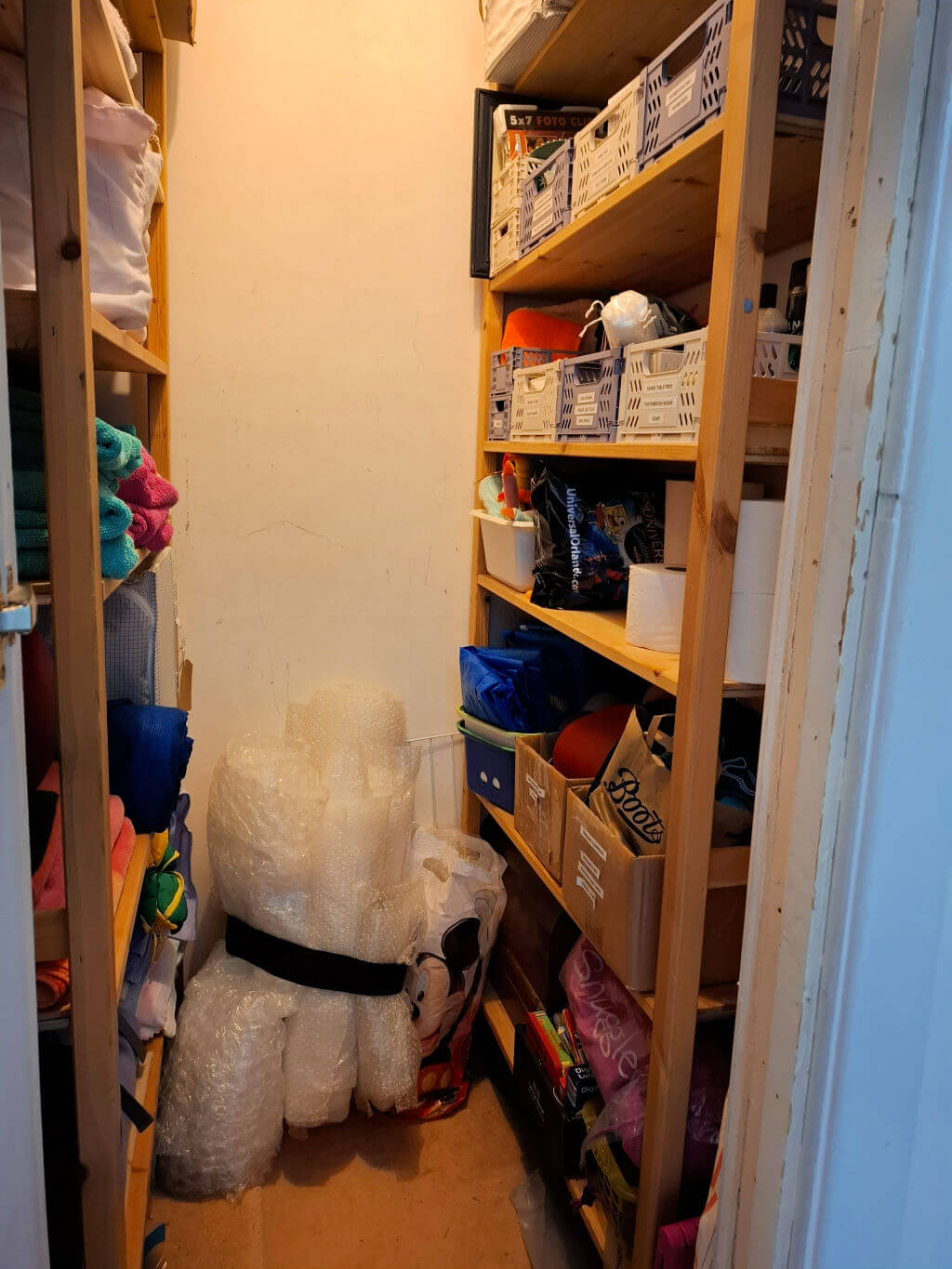 A small storage room filled with wooden shelves lined with plastic bins and assorted items. Rolls of bubble wrap are stacked against a wall, creating an organised space.