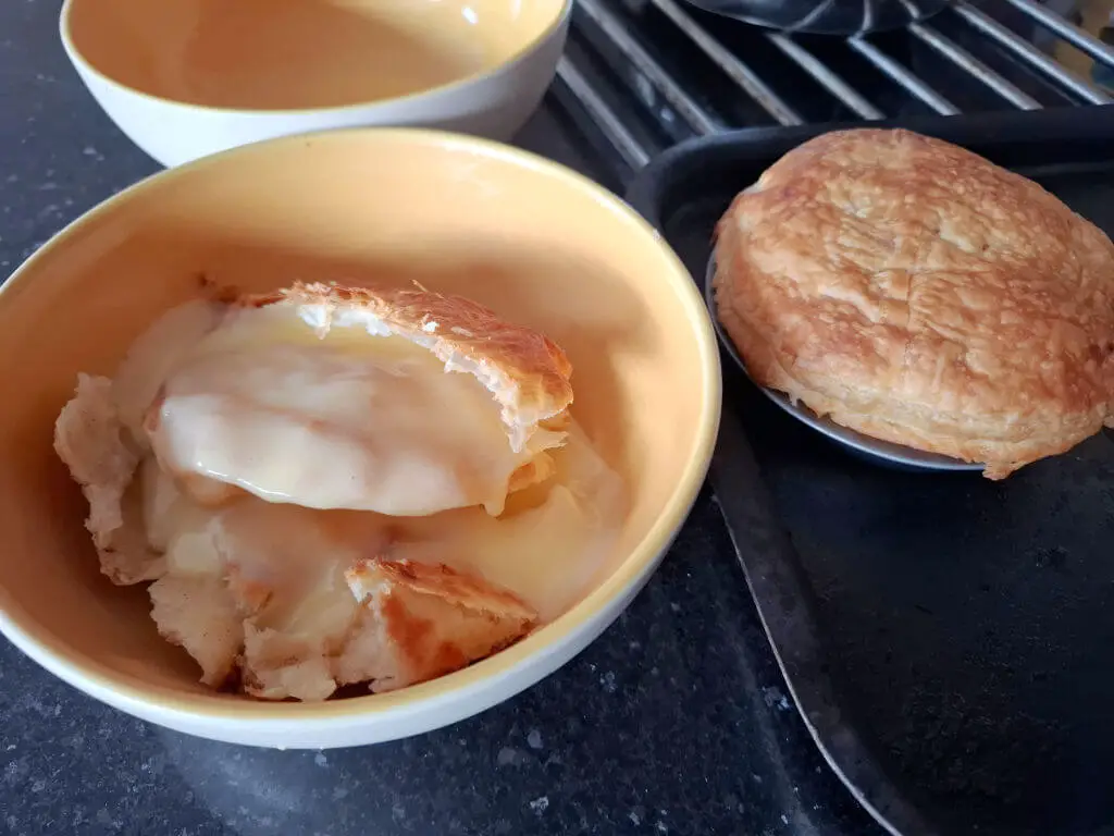 Bowl with broken pie showing creamy filling beside whole pie on tray. Warm, inviting tones with golden pastry. Cosy and appetizing atmosphere.
