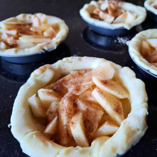 Five mini pie crusts filled with sliced apples, sprinkled with cinnamon, on a dark countertop. The image evokes a cozy, homemade feel.