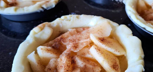 Five mini pie crusts filled with sliced apples, sprinkled with cinnamon, on a dark countertop. The image evokes a cozy, homemade feel.