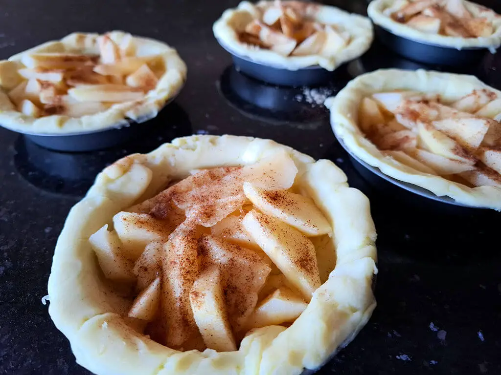 Five mini pie crusts filled with sliced apples, sprinkled with cinnamon, on a dark countertop. The image evokes a cozy, homemade feel.