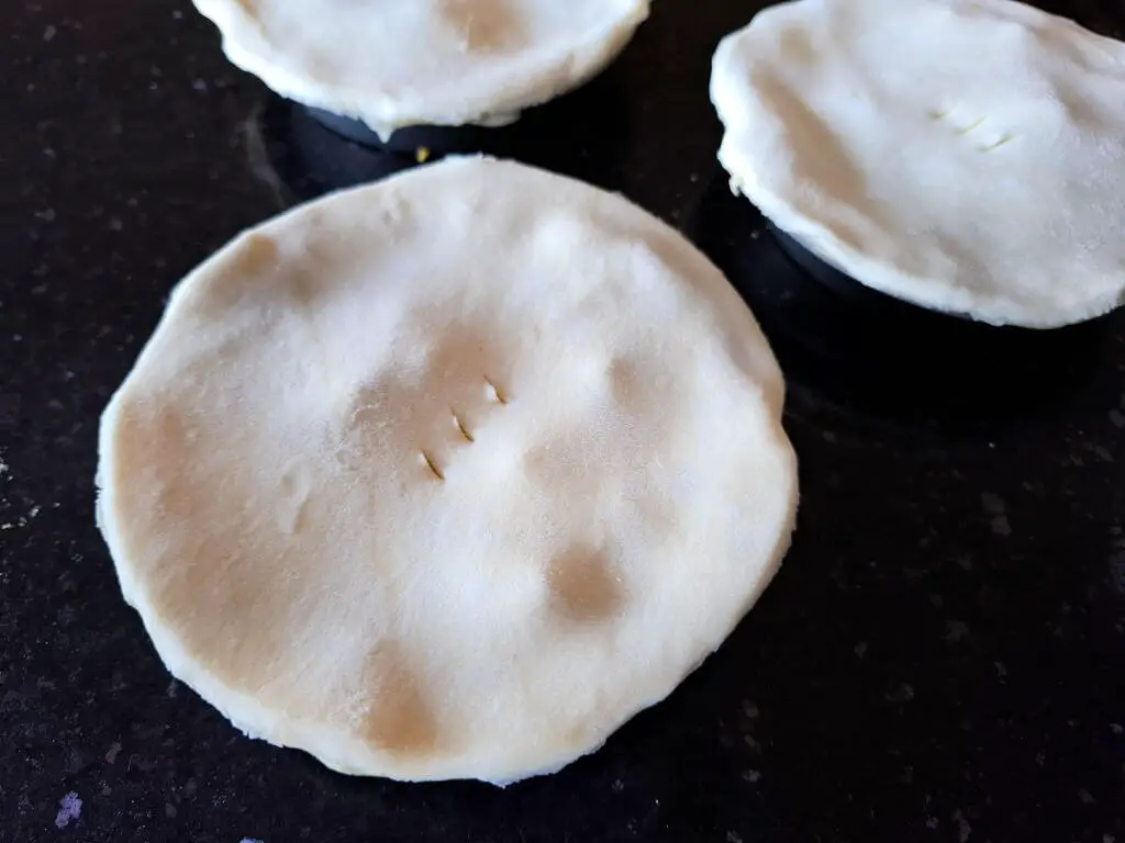 Three round, uncooked pies with smooth pastry tops and three small slits each are placed on a dark countertop, ready for baking.