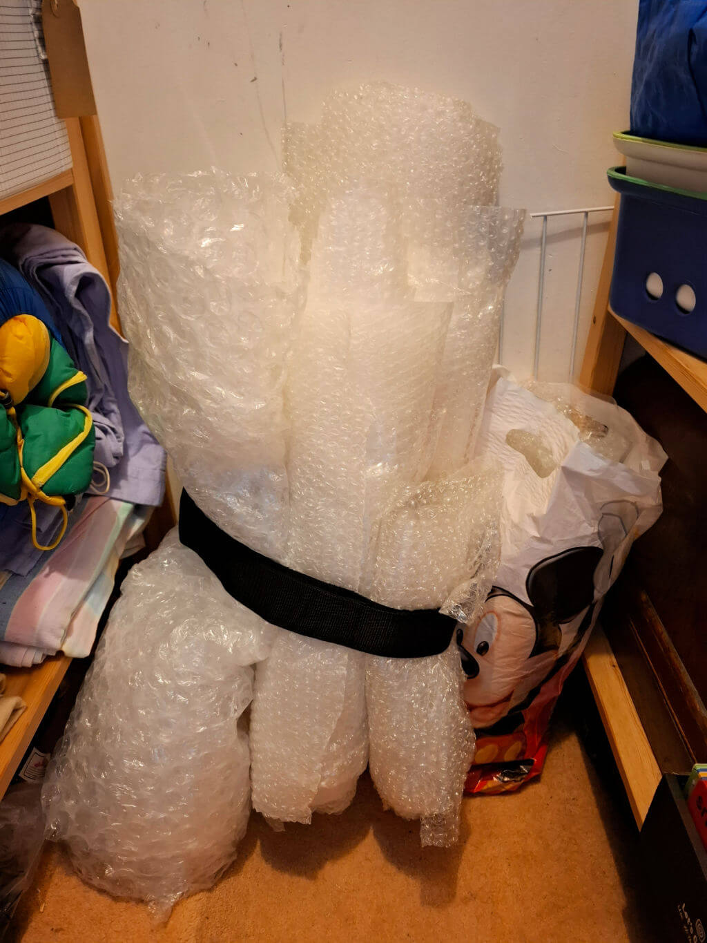 Rolls of bubble wrap secured with a black strap stand against a wall in a cluttered room. Nearby are colourful bags and folded clothes, evoking a storage space.