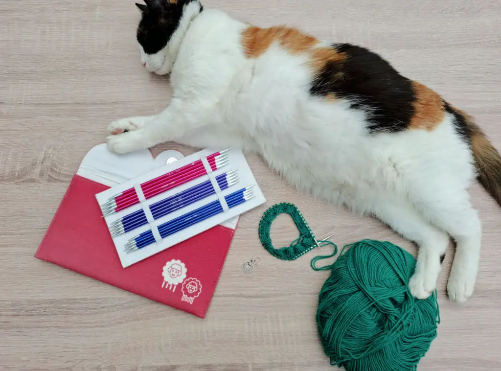 Cat sleeping on wooden floor beside knitting needles, green yarn, and a partially knit sock. Calm and cozy crafting scene.