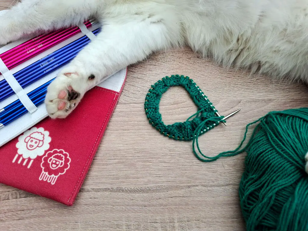 A fluffy white cat's paws rest on a red and white knitting needle case. Nearby, green yarn and a knitting project suggest a cozy crafting scene.
