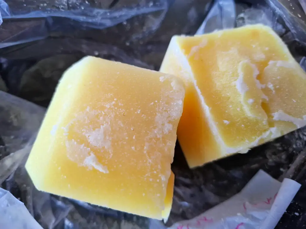 Two rectangular chunks of frozen yellow sauce rest on a partially unwrapped plastic packaging. The surface shows frost.