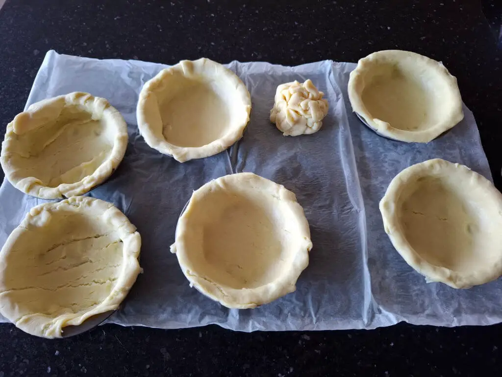 Six unbaked pastry shells and a dough ball sit on parchment paper on a dark countertop, ready for baking. The scene conveys a sense of anticipation.