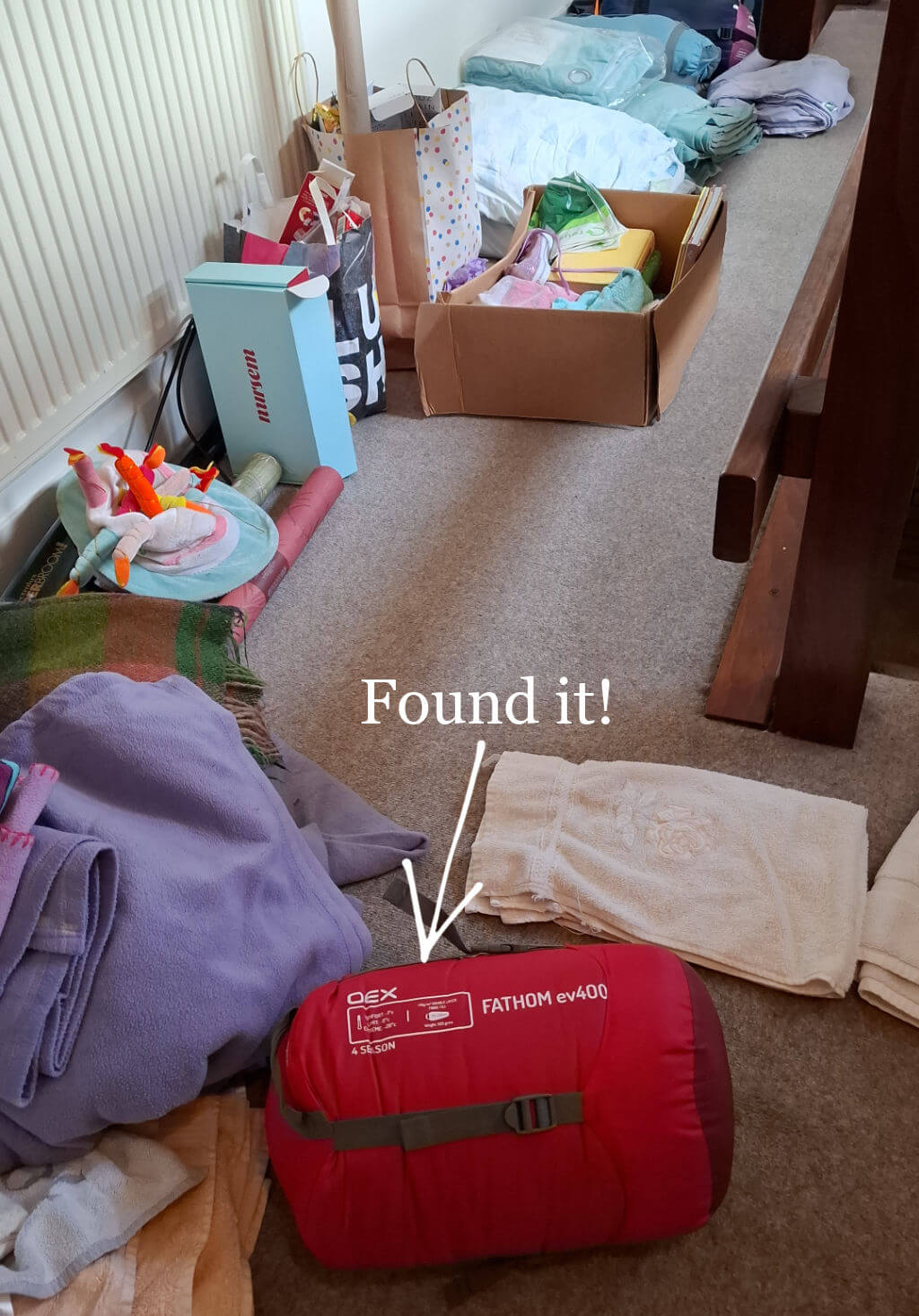 A cluttered room with stacked blankets, towels, boxes, and bags. A red sleeping bag with "Found it!" text and arrow highlights its significance.