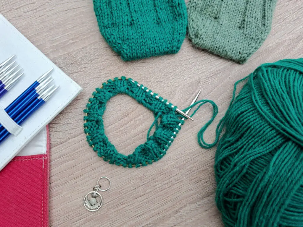 Knitting scene with green yarn, circular needles on a project, completed green and mint socks, blue knitting needles in a case, and a stitch marker on a wooden surface.