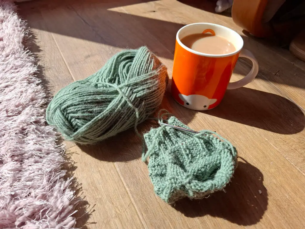 A ball of green yarn and a partially knitted sock sit on a wooden floor with sunlight. A bright orange mug with a sheep face holds tea nearby.