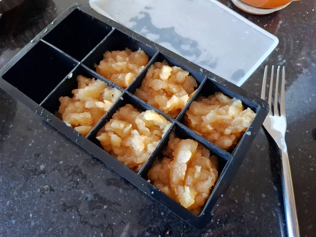 Frozen apple sauce cubes in a black, six-compartment ice tray on a dark countertop, accompanied by a fork, conveying a practical and casual tone.