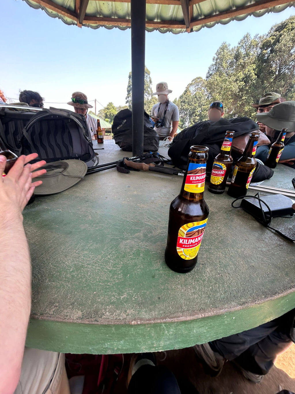 A group of people sitting around a circular wooden table under a canopy, with backpacks and Kilimanjaro beer bottles. The setting is outdoors, surrounded by trees.