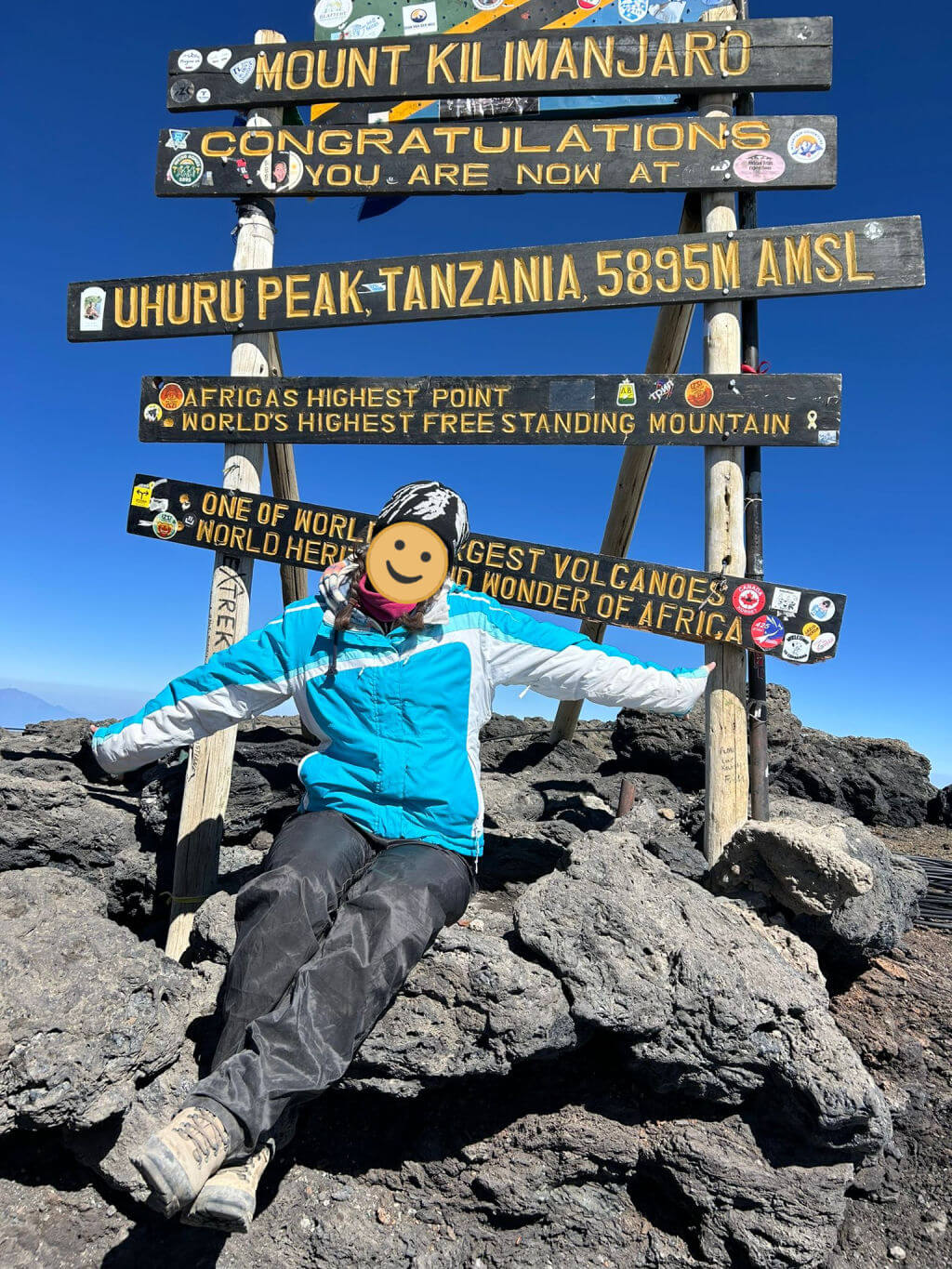 Not so small daughter in hiking gear joyfully poses on rocky terrain at Uhuru Peak, Mount Kilimanjaro, next to a colourful sign marking Africa's highest point.