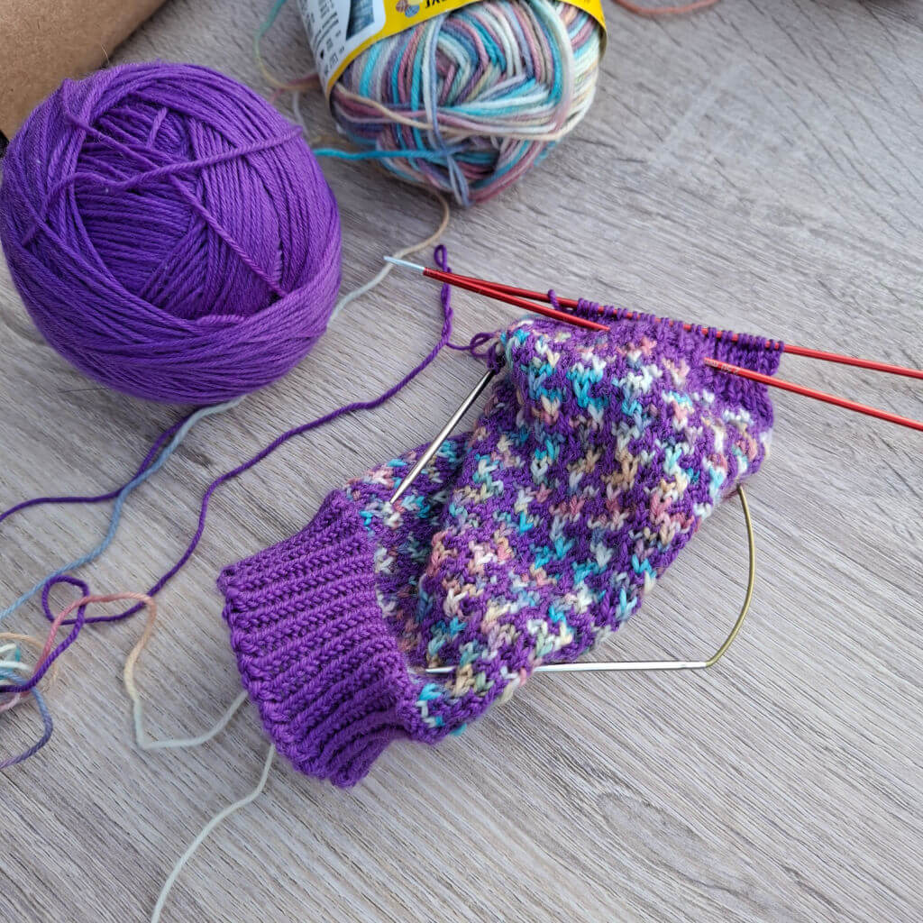 Purple and multicoloured knitting project on wooden surface, with red double-pointed needles and two yarn balls, one purple and one pastel. Cosy and creative.