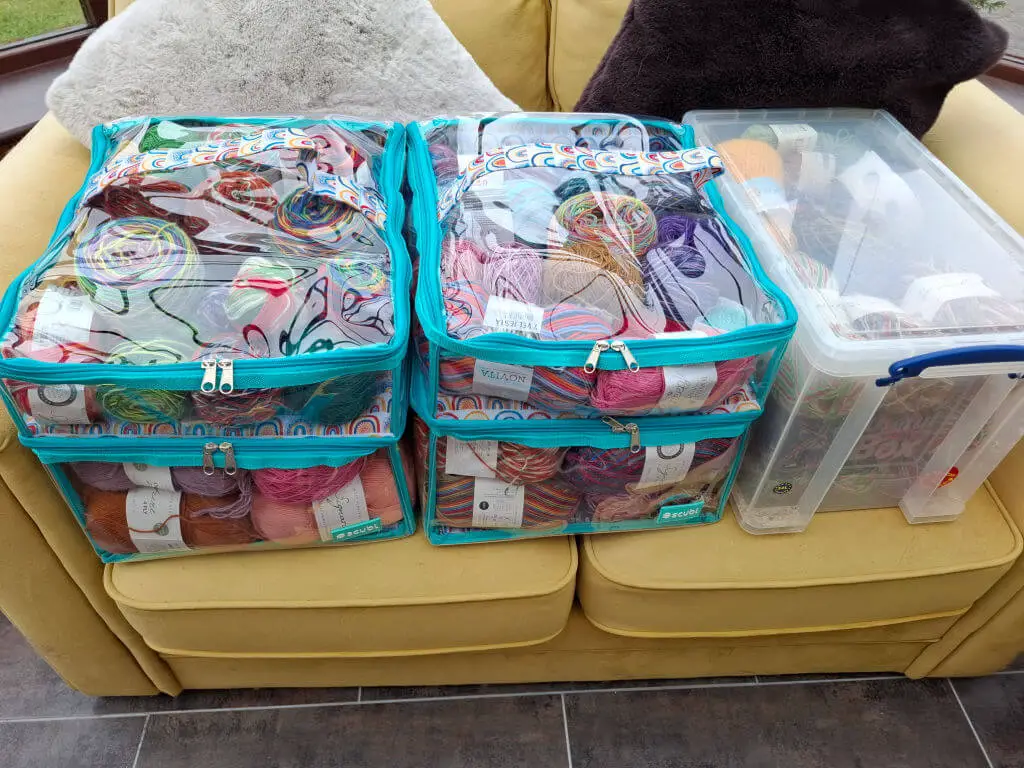 Two clear storage bags and a box filled with colourful yarns are arranged on a yellow sofa. The scene exudes creativity and organisation.