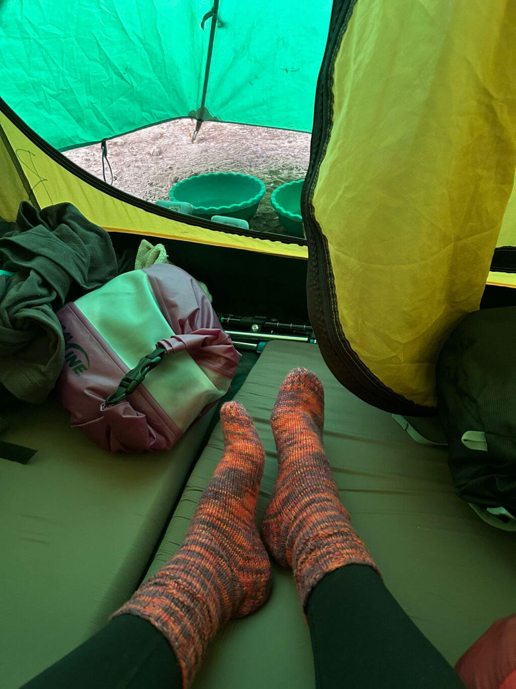View from inside a tent with two feet in orange socks resting on a sleeping pad. A pink backpack and green bowls are visible, evoking a cozy camping vibe.