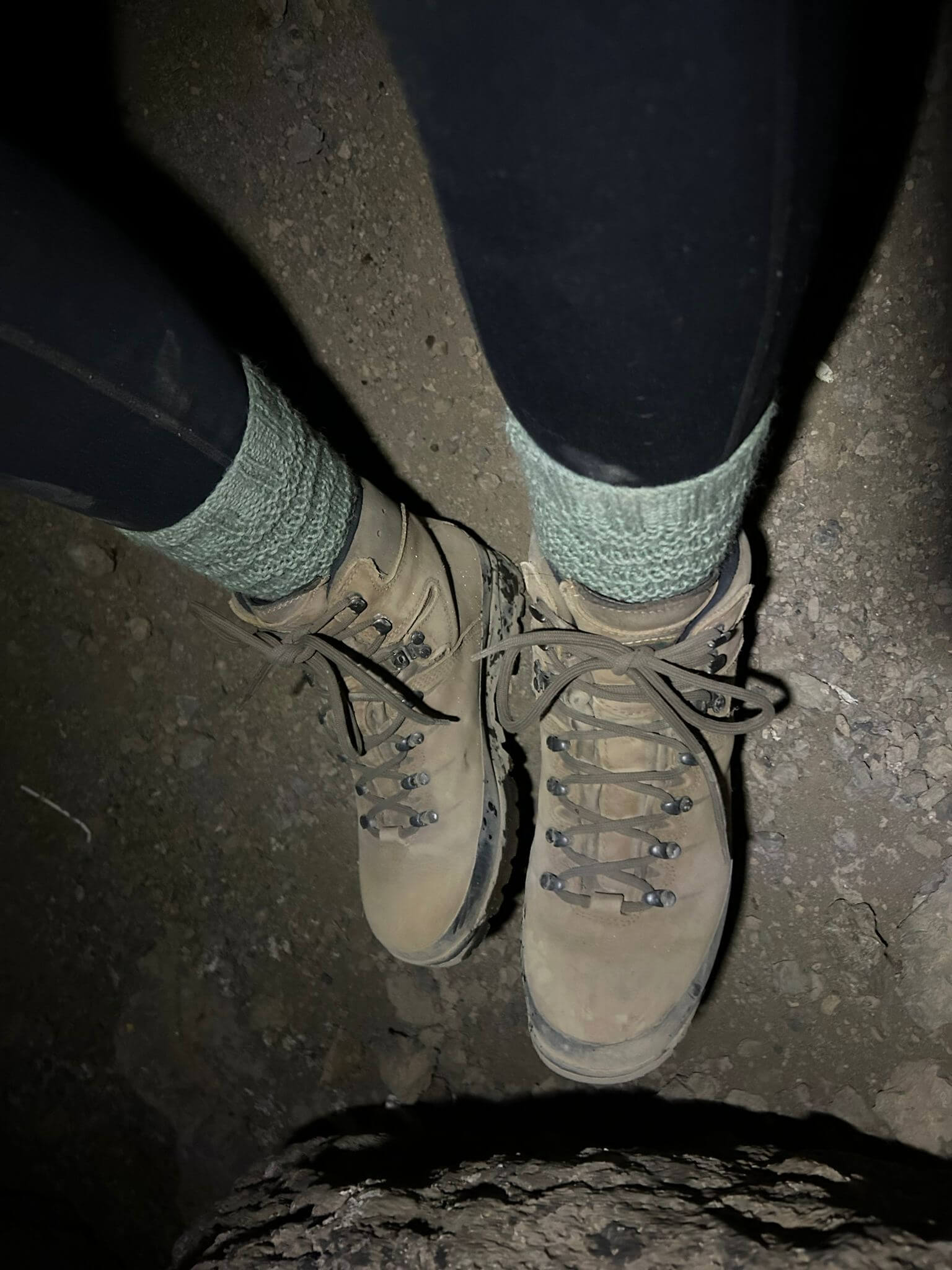 A person wearing tan hiking boots and green socks stands on a rocky, dirt path. The dim lighting suggests a nighttime or early morning trek.