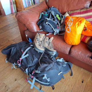 Backpacks, hiking boots, and an orange bag sit on a brown leather sofa and wooden floor.