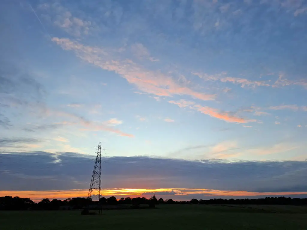 Sunset over a countryside landscape with a silhouetted pylon. The sky features soft pink and blue hues, creating a serene atmosphere.