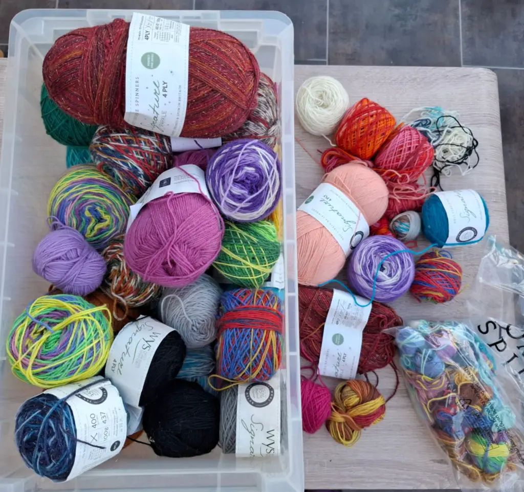 Various colourful yarn balls are organized in and around a transparent plastic container on a wooden surface. The scene conveys creativity and warmth.