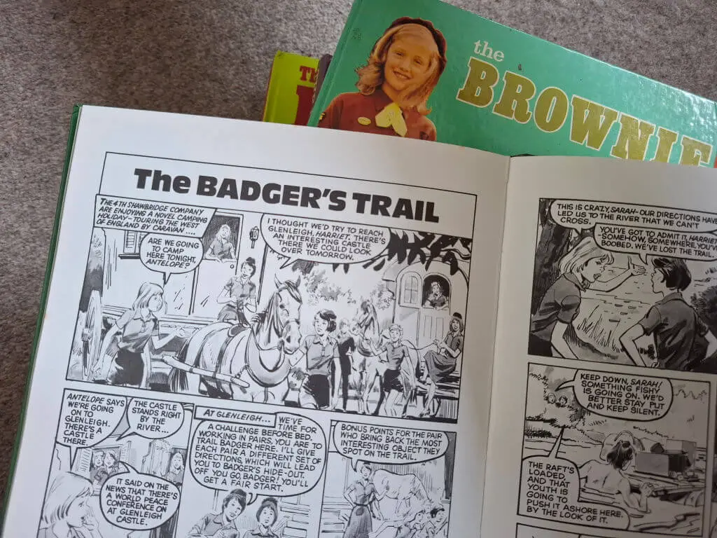 Open Brownie annual book with a story titled "The Badger's Trail" shows black-and-white illustrations of characters on a horse-drawn cart. Background includes a green "The Brownie" book.