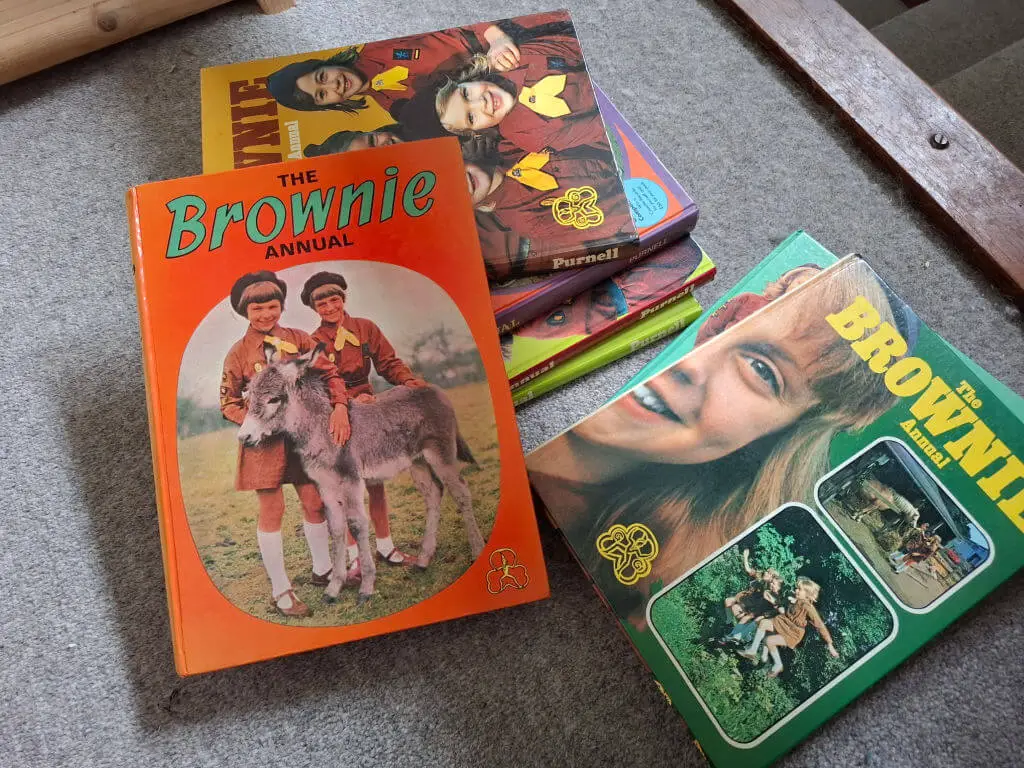 Stack of vintage "The Brownie Annual" books on a grey carpet. Prominent orange cover shows children in uniforms with a donkey, evoking nostalgia.