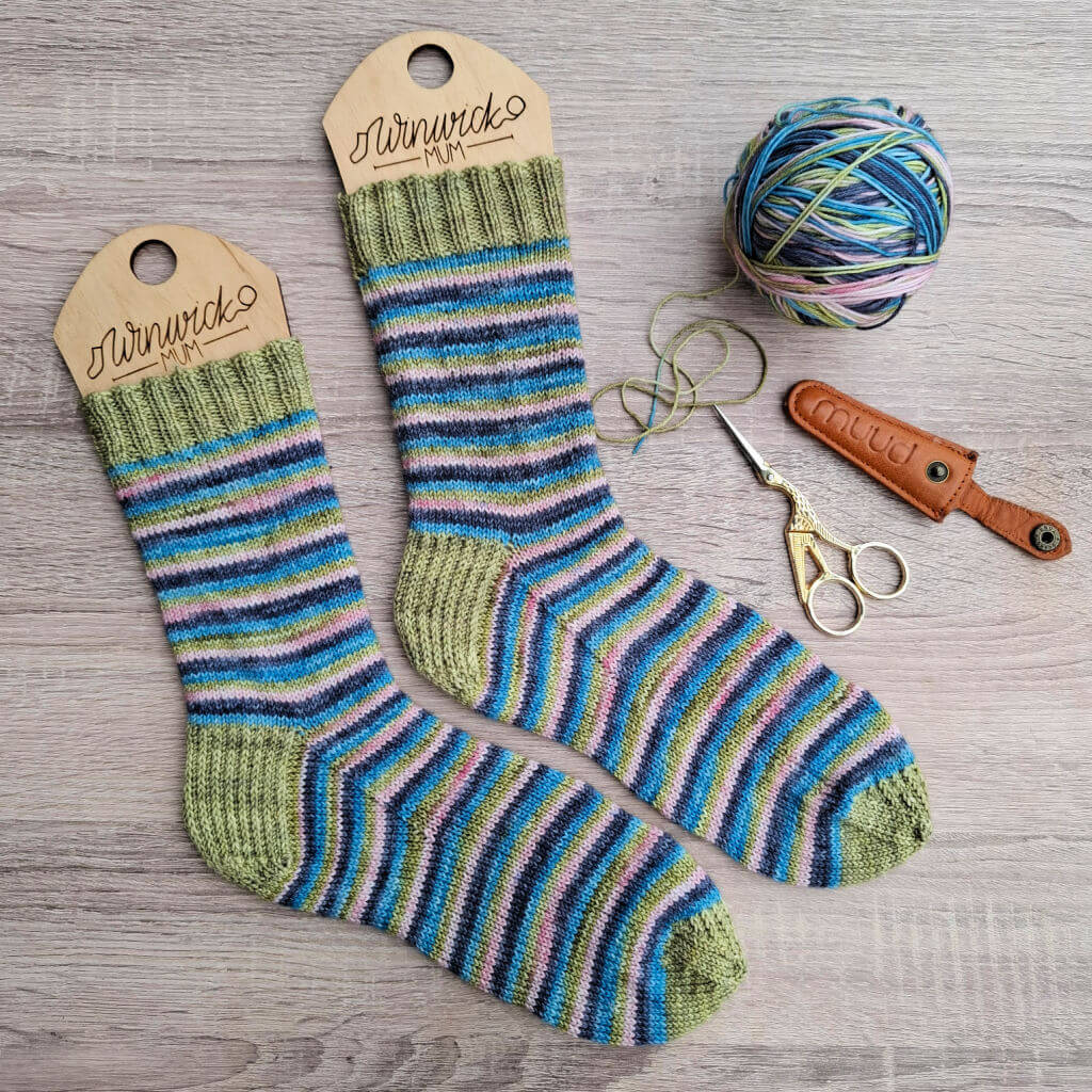 Striped knitted socks in shades of green, blue, and purple on wooden sock blockers. Beside them, matching yarn, scissors, and a tan leather cover lie on a wooden surface.