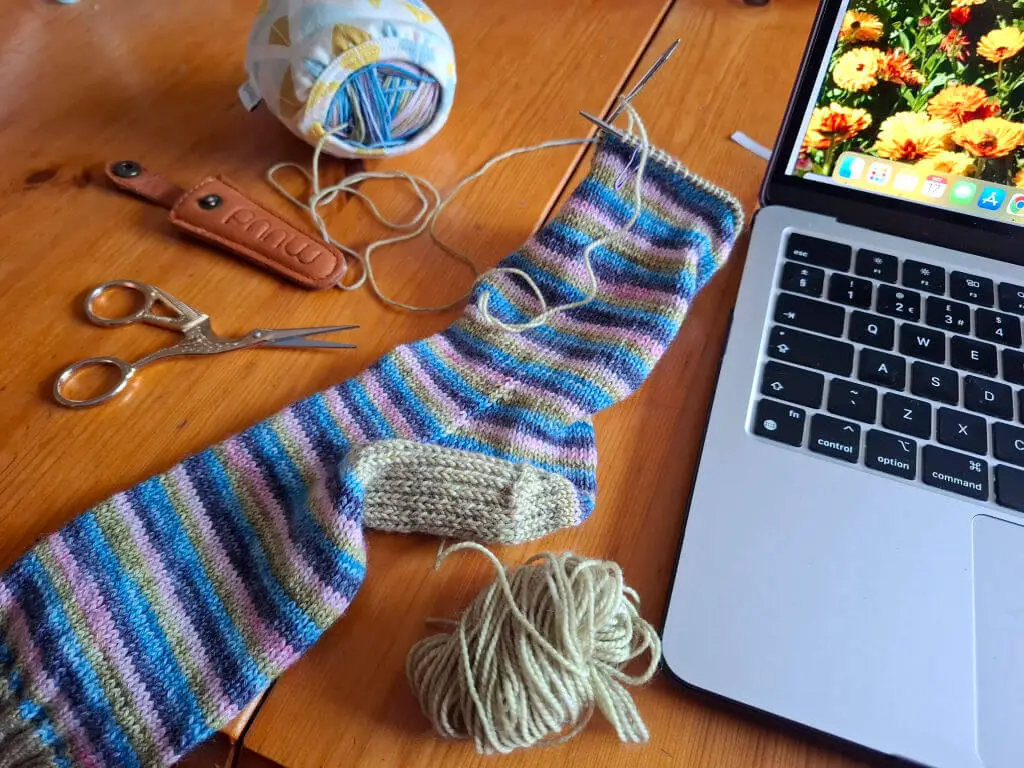 Knitting project with colourful striped sock, yarn, and needles on a wooden table next to a laptop. Scissors and knit bag add a cosy, creative vibe.