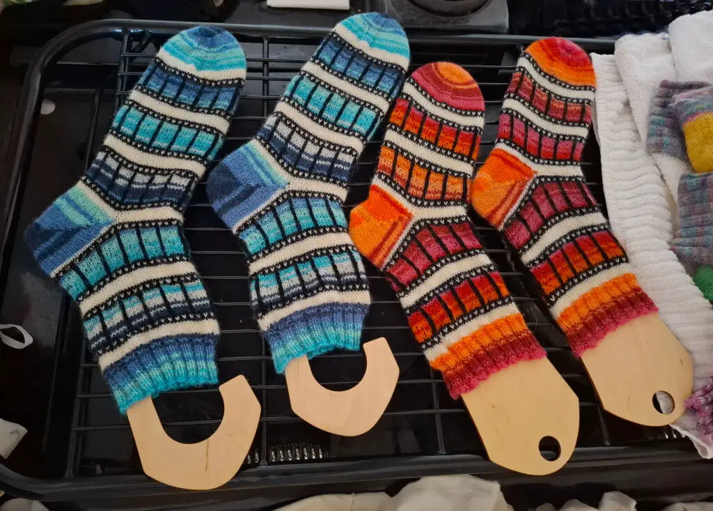 Four colourful, patterned socks drying on wooden blockers on a metal rack. Two are in blue hues, and two in red-orange tones with a film-reel pattern
