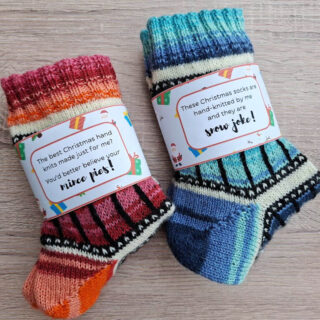 Two pairs of colourful, hand-knit Christmas socks on a wooden surface. Each has a festive label with playful holiday messages, creating a cheerful vibe.