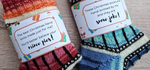 Two pairs of colourful, hand-knit Christmas socks on a wooden surface. Each has a festive label with playful holiday messages, creating a cheerful vibe.