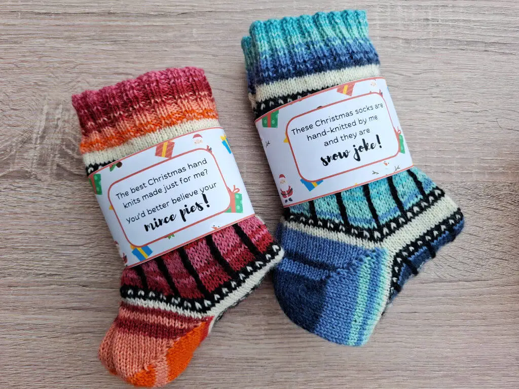 Two pairs of colourful, hand-knit Christmas socks on a wooden surface. Each has a festive label with playful holiday messages, creating a cheerful vibe.