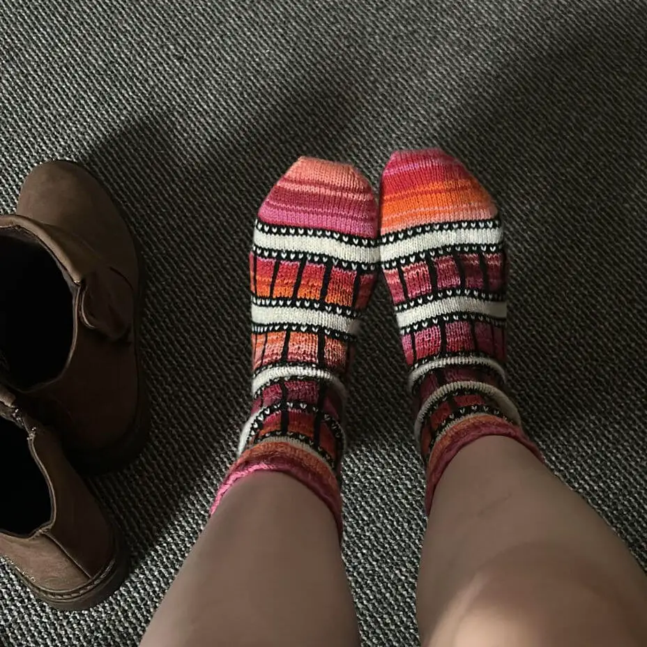 Feet in colourful, patterned socks with a film-reel design on a textured grey carpet, next to a pair of brown boots, conveying warmth and cosiness.