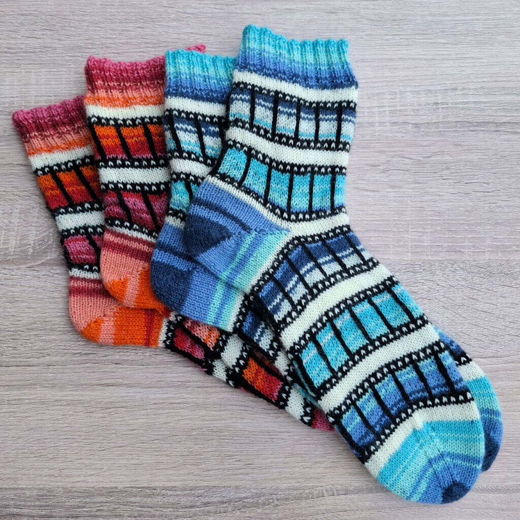 Colourful socks with a film-reel pattern are neatly stacked on a light wooden surface. The socks feature vibrant shades of blue, pink, orange, and black.