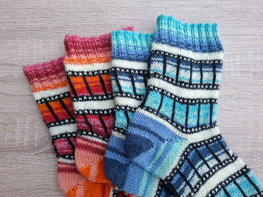 Four colourful knit socks with a colourwork film reel pattern are fanned out on a wooden surface. The socks display shades of orange, pink, teal, and blue.