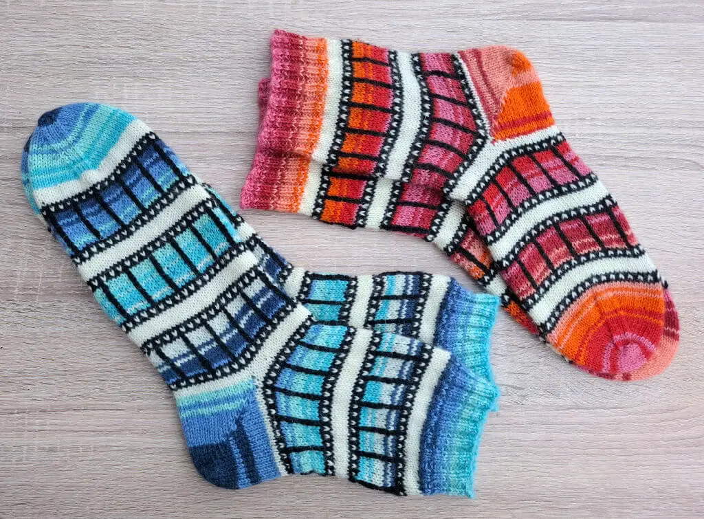 Colourful knitted socks on a wooden surface. One pair has a blue and teal pattern; the other features red and orange hues, both with a film-reel design.