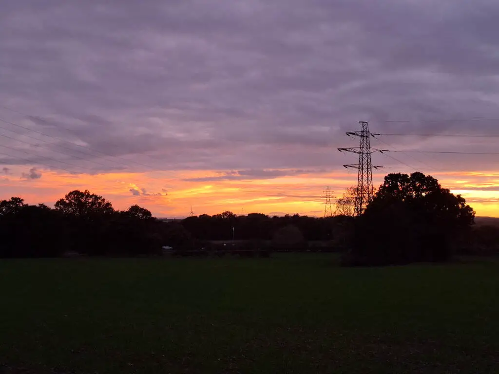 Sunset over a field, with orange and purple hues in the sky. Silhouetted trees and power lines create a serene yet industrial contrast.