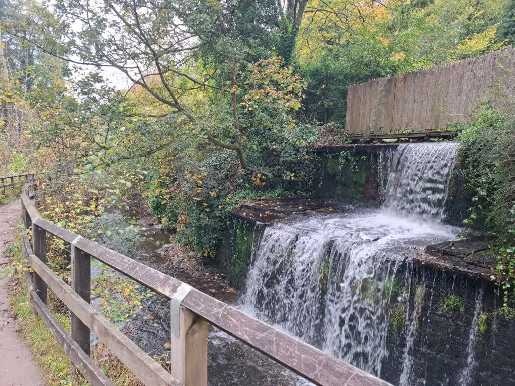 A tranquil waterfall cascades over mossy rocks surrounded by lush greenery. A wooden fence line and trail run alongside, hinting at a peaceful hike.